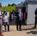 A group of approximately 10-12 professionals in business attire are gathered outdoors at what appears to be a facility tour or site visit. A central figure in a gray suit and blue tie is actively speaking and gesturing, suggesting he is leading the tour or presentation. The setting includes industrial or utility equipment enclosures, gravel landscaping, and a manicured green lawn in the background.