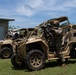 Military personnel in full combat gear are conducting what appears to be a training exercise using tan/desert-colored tactical utility vehicles (UTVs). At least two vehicles are visible, with soldiers positioned in and around them, one actively aiming a rifle from the vehicle. The setting is an outdoor training area with a concrete block building and vegetation in the background.