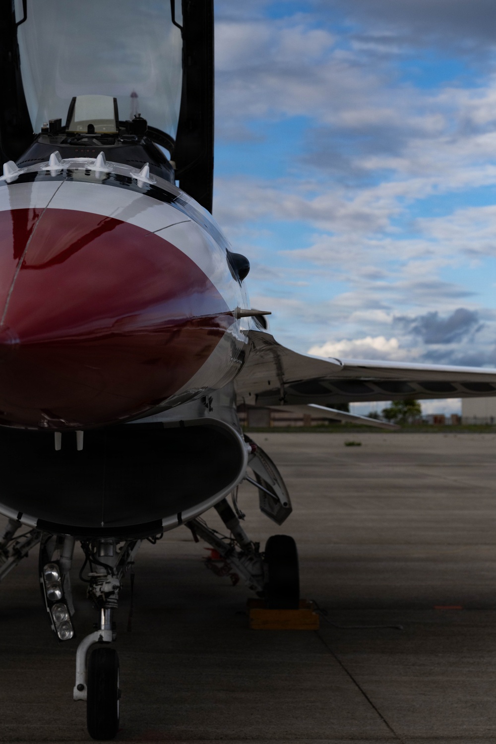 A close-up ground-level photograph of a military jet fighter, likely an F-16 Fighting Falcon, featuring a distinctive red and white paint scheme consistent with the USAF Thunderbirds demonstration team. The aircraft is parked on a concrete flight line or tarmac, with the nose, cockpit canopy, forward landing gear, and wing root visible. The image is taken from a low angle emphasizing the aircraft's fuselage and underside details against a partly cloudy sky.