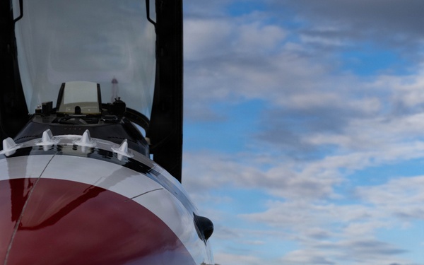 A close-up ground-level photograph of a military jet fighter, likely an F-16 Fighting Falcon, featuring a distinctive red and white paint scheme consistent with the USAF Thunderbirds demonstration team. The aircraft is parked on a concrete flight line or tarmac, with the nose, cockpit canopy, forward landing gear, and wing root visible. The image is taken from a low angle emphasizing the aircraft's fuselage and underside details against a partly cloudy sky.