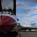 A close-up ground-level photograph of a military jet fighter, likely an F-16 Fighting Falcon, featuring a distinctive red and white paint scheme consistent with the USAF Thunderbirds demonstration team. The aircraft is parked on a concrete flight line or tarmac, with the nose, cockpit canopy, forward landing gear, and wing root visible. The image is taken from a low angle emphasizing the aircraft's fuselage and underside details against a partly cloudy sky.