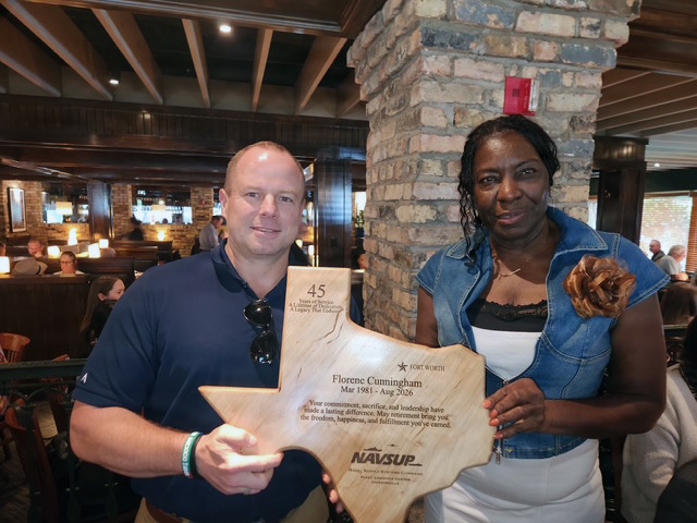 Two individuals are posing together in a restaurant setting, holding a wooden plaque shaped like the state of Texas. The plaque is a retirement award presented by NAVSUP (Naval Supply Systems Command) to Florene Cunningham, recognizing 45 years of service from March 1981 to August 2026. The event appears to be a retirement celebration held at a casual dining establishment.
