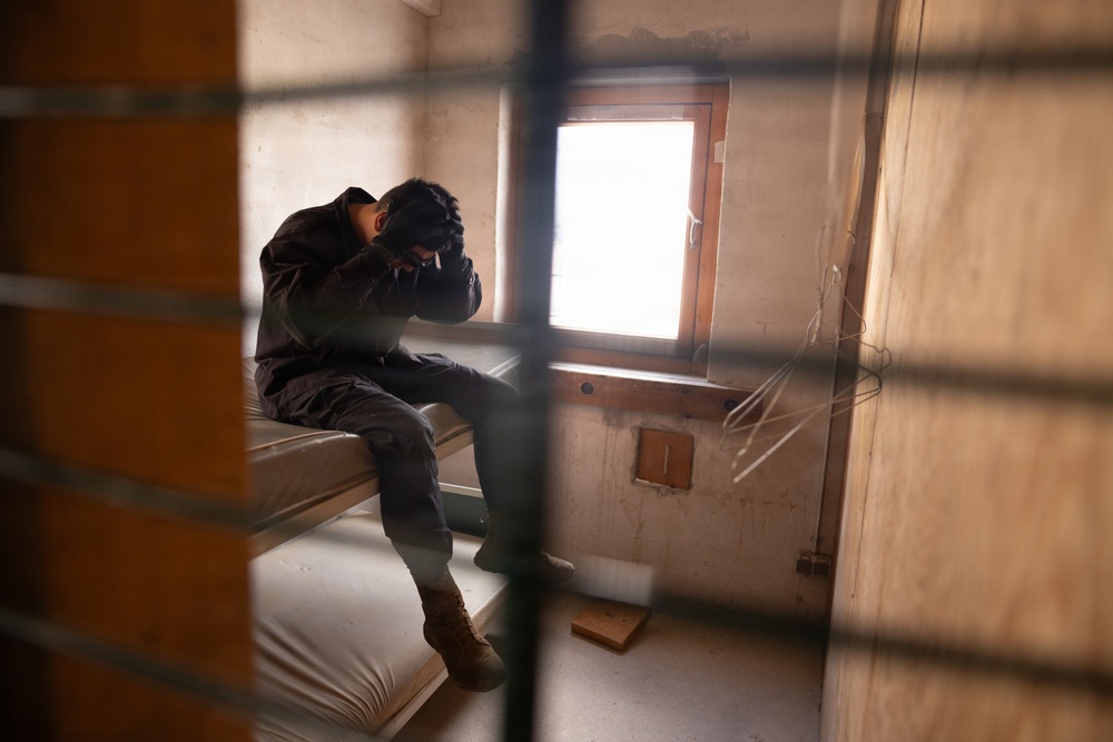 A person is seated on a bare mattress inside a sparse, confined room, viewed through what appears to be bars or a grated barrier in the foreground. The individual has their head bowed and hands covering their face, suggesting distress or despair. The room contains minimal furnishings and has a small window providing the primary light source.