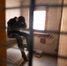 A person is seated on a bare mattress inside a sparse, confined room, viewed through what appears to be bars or a grated barrier in the foreground. The individual has their head bowed and hands covering their face, suggesting distress or despair. The room contains minimal furnishings and has a small window providing the primary light source.
