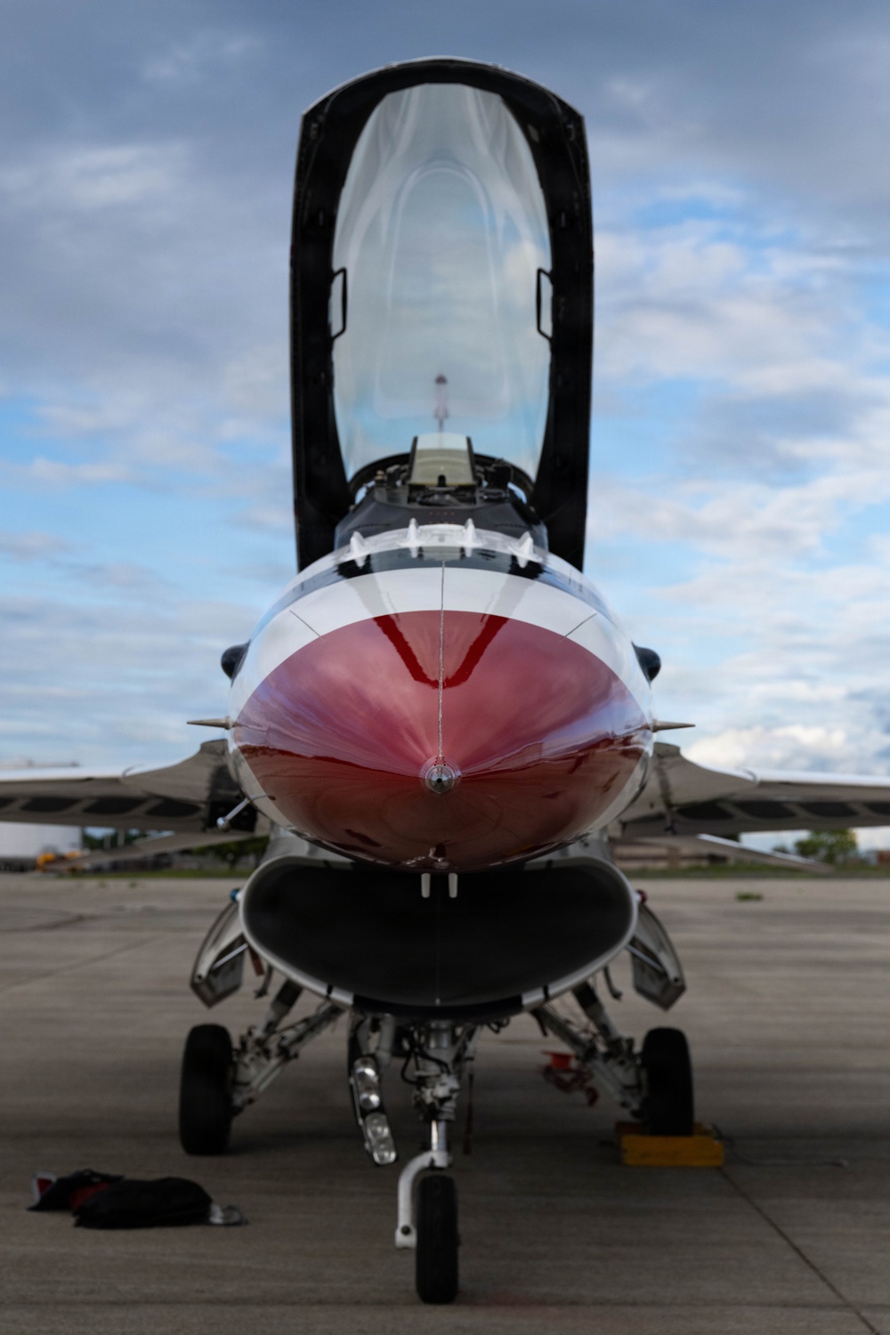A front-facing close-up photograph of a U.S. Air Force Thunderbirds F-16 Fighting Falcon parked on a tarmac apron. The aircraft's canopy is open and raised, revealing the empty cockpit interior. The distinctive red and white Thunderbirds livery is prominently visible on the nose cone and fuselage.