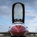A front-facing close-up photograph of a U.S. Air Force Thunderbirds F-16 Fighting Falcon parked on a tarmac apron. The aircraft's canopy is open and raised, revealing the empty cockpit interior. The distinctive red and white Thunderbirds livery is prominently visible on the nose cone and fuselage.