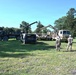 Military personnel are conducting a vehicle recovery or lifting operation in an open grassy field, using at least two large military trucks equipped with crane/boom arms. Three soldiers wearing camouflage uniforms and white hard hats are observing or supervising the operation. The scene appears to be a training exercise or field maintenance operation at a military installation.