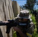 Two armed individuals in camouflage uniforms are conducting what appears to be a military training exercise in an outdoor environment. The foreground figure is in a crouched tactical position holding a rifle against a concrete or panel barrier, while a second figure stands in the background holding a weapon. Both wear multicam-pattern uniforms with yellow identification markings on their arms.