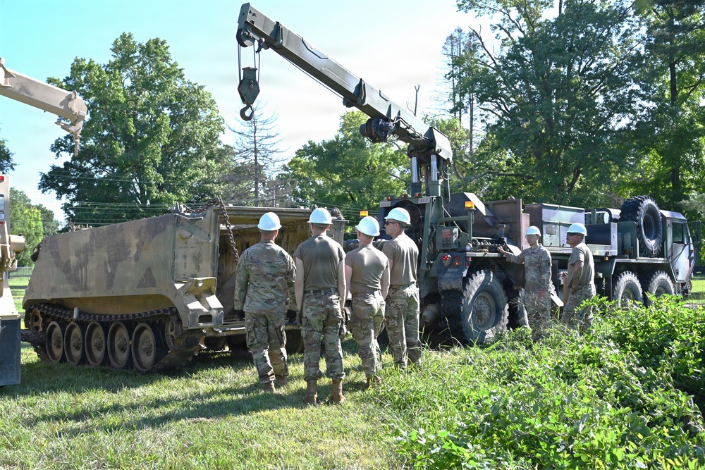 A group of military personnel wearing OCP (Operational Camouflage Pattern) uniforms and white hard hats are gathered around military vehicles in a grassy outdoor area. A large military crane vehicle (appears to be an M984 HEMTT wrecker or similar recovery vehicle) is positioned next to a tracked armored vehicle, suggesting a vehicle recovery or maintenance training exercise. The scene appears to be a training environment given the instructional grouping of soldiers observing the operation.