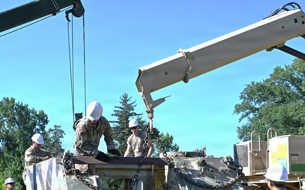 Military personnel in camouflage uniforms and white hard hats are working to recover or dismantle a heavily damaged tracked armored vehicle, likely an M113 armored personnel carrier. Two crane vehicles are being used simultaneously to lift components from the wrecked vehicle. The operation appears to be a recovery or salvage exercise conducted in an outdoor grassy area near tree lines.