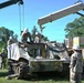 Military personnel in camouflage uniforms and white hard hats are working to recover or dismantle a heavily damaged tracked armored vehicle, likely an M113 armored personnel carrier. Two crane vehicles are being used simultaneously to lift components from the wrecked vehicle. The operation appears to be a recovery or salvage exercise conducted in an outdoor grassy area near tree lines.