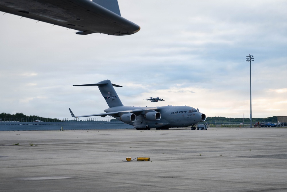 A U.S. Air Force C-17 Globemaster III transport aircraft is parked on a military flight line apron. The aircraft bears the markings 'U.S. AIR FORCE' and the serial number '0057', with 'STEWART' visible on the tail, indicating assignment to Stewart Air National Guard Base. A partial wing of another aircraft is visible in the foreground upper left, and a second aircraft appears to be airborne in the background.