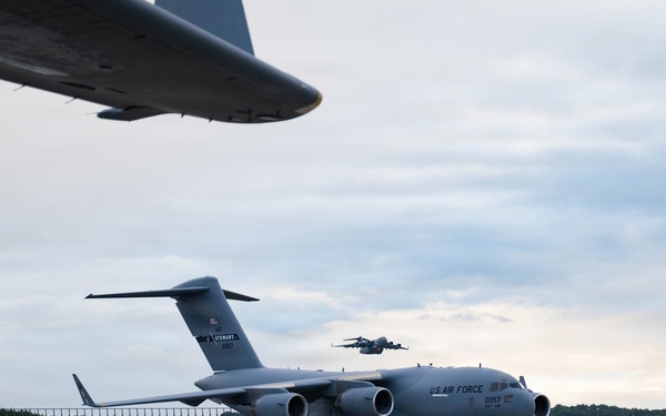 A U.S. Air Force C-17 Globemaster III transport aircraft is parked on a military flight line apron. The aircraft bears the markings 'U.S. AIR FORCE' and the serial number '0057', with 'STEWART' visible on the tail, indicating assignment to Stewart Air National Guard Base. A partial wing of another aircraft is visible in the foreground upper left, and a second aircraft appears to be airborne in the background.