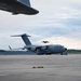 A U.S. Air Force C-17 Globemaster III transport aircraft is parked on a military flight line apron. The aircraft bears the markings 'U.S. AIR FORCE' and the serial number '0057', with 'STEWART' visible on the tail, indicating assignment to Stewart Air National Guard Base. A partial wing of another aircraft is visible in the foreground upper left, and a second aircraft appears to be airborne in the background.