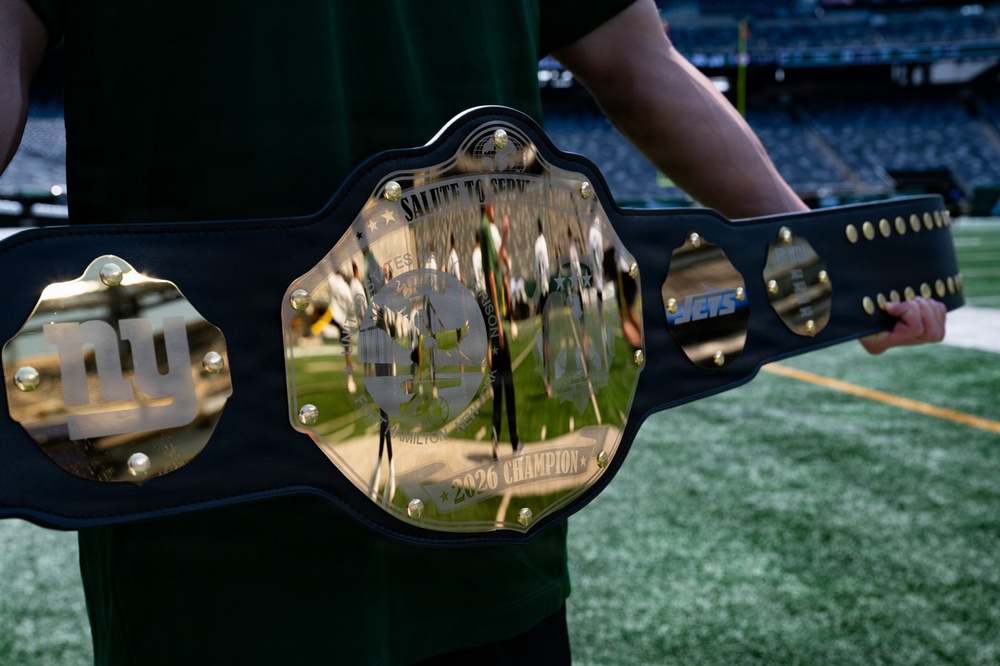 A large decorative championship-style belt is being held by a person on what appears to be an NFL football field. The belt features gold plates with 'Salute to Service' text, '2026 Champion' inscription, and logos from the New York Giants (NY) and New York Jets on the side plates. The setting appears to be MetLife Stadium, home of both New York NFL teams.