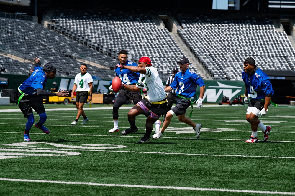 A flag football game is being played on the field of MetLife Stadium, home of the New York Jets, as indicated by the Jets logo visible on the stadium wall. Two teams — one in blue jerseys and one in green/white jerseys — are actively engaged in a play, with a ball carrier in a white jersey and red bandana being pursued by defenders. The stadium seats are empty, suggesting this is a non-game-day event such as a charity game, promotional event, or community activity.