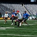 A flag football game is being played on the field of MetLife Stadium, home of the New York Jets, as indicated by the Jets logo visible on the stadium wall. Two teams — one in blue jerseys and one in green/white jerseys — are actively engaged in a play, with a ball carrier in a white jersey and red bandana being pursued by defenders. The stadium seats are empty, suggesting this is a non-game-day event such as a charity game, promotional event, or community activity.
