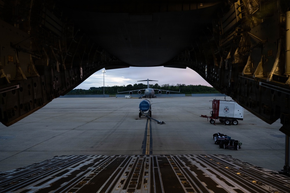 The image is taken from inside the open cargo bay of a large military transport aircraft, looking outward onto a flight line apron at dusk or dawn. A second large military transport aircraft is visible in the background on the tarmac, along with ground support equipment. The perspective through the cargo ramp creates a dramatic framing effect with the exterior scene.