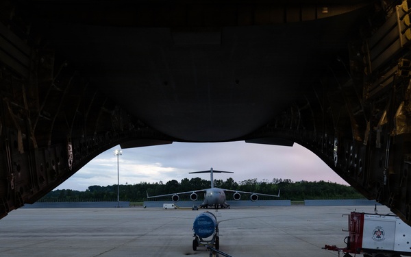 The image is taken from inside the open cargo bay of a large military transport aircraft, looking outward onto a flight line apron at dusk or dawn. A second large military transport aircraft is visible in the background on the tarmac, along with ground support equipment. The perspective through the cargo ramp creates a dramatic framing effect with the exterior scene.