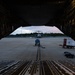 The image is taken from inside the open cargo bay of a large military transport aircraft, looking outward onto a flight line apron at dusk or dawn. A second large military transport aircraft is visible in the background on the tarmac, along with ground support equipment. The perspective through the cargo ramp creates a dramatic framing effect with the exterior scene.