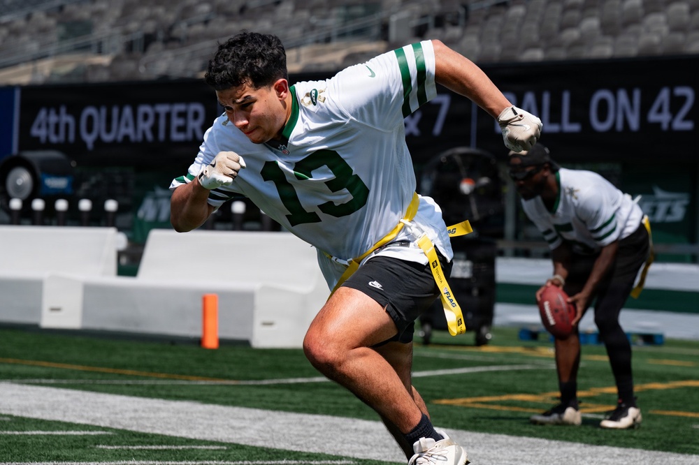 A young athlete wearing jersey number 13 in white and green is playing flag football on a professional NFL field, identifiable as a New York Jets stadium. The player is in an aggressive running stance with yellow flag belts attached at the waist. A second player is visible in the background holding a football in a crouched position.