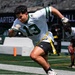 A young athlete wearing jersey number 13 in white and green is playing flag football on a professional NFL field, identifiable as a New York Jets stadium. The player is in an aggressive running stance with yellow flag belts attached at the waist. A second player is visible in the background holding a football in a crouched position.
