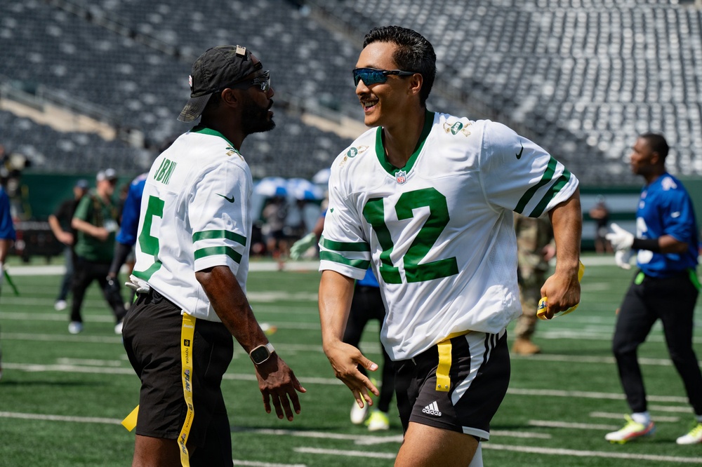 Two men wearing New York Jets NFL jerseys (numbers 5 and 12) are laughing and interacting on a football field during what appears to be a flag football event. Both are wearing yellow flag football belts and sunglasses, suggesting a casual or charity game setting. The stadium appears largely empty with a few other participants visible in the background.