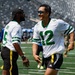 Two men wearing New York Jets NFL jerseys (numbers 5 and 12) are laughing and interacting on a football field during what appears to be a flag football event. Both are wearing yellow flag football belts and sunglasses, suggesting a casual or charity game setting. The stadium appears largely empty with a few other participants visible in the background.