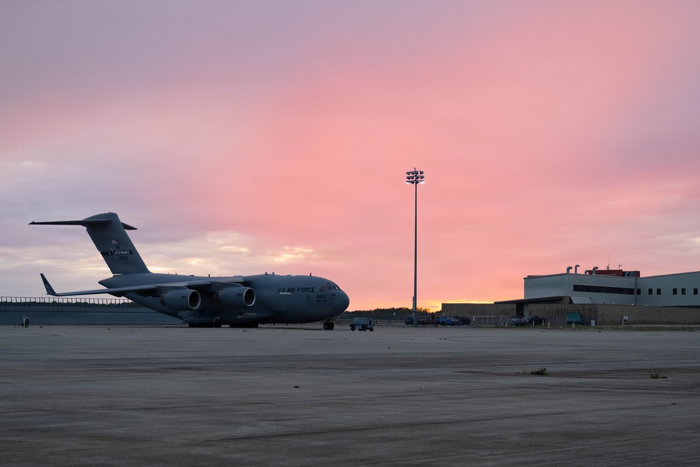 A U.S. Air Force C-17 Globemaster III military transport aircraft is parked on a flight line apron at dusk or dawn, with a vivid pink and purple sky in the background. The aircraft is positioned on the left side of the frame, with military hangars and a tall floodlight tower visible to the right. The scene appears to be at a U.S. Air Force base or military airfield.