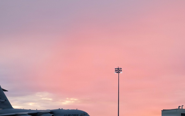 A U.S. Air Force C-17 Globemaster III military transport aircraft is parked on a flight line apron at dusk or dawn, with a vivid pink and purple sky in the background. The aircraft is positioned on the left side of the frame, with military hangars and a tall floodlight tower visible to the right. The scene appears to be at a U.S. Air Force base or military airfield.