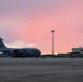 A U.S. Air Force C-17 Globemaster III military transport aircraft is parked on a flight line apron at dusk or dawn, with a vivid pink and purple sky in the background. The aircraft is positioned on the left side of the frame, with military hangars and a tall floodlight tower visible to the right. The scene appears to be at a U.S. Air Force base or military airfield.