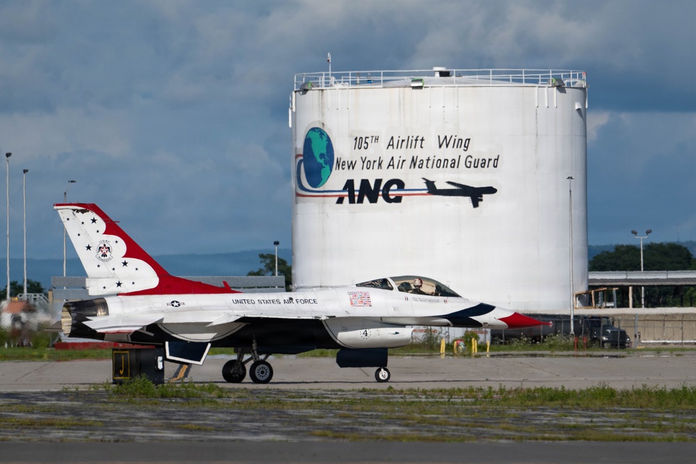 A United States Air Force Thunderbirds F-16 Fighting Falcon, numbered '4', is taxiing on the tarmac at Stewart Air National Guard Base. The aircraft displays the iconic red, white, and blue Thunderbirds livery. In the background, a large fuel storage tank bears the markings of the 105th Airlift Wing, New York Air National Guard (ANG).