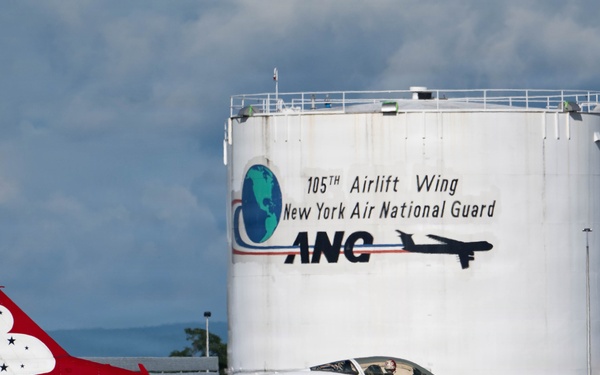 A United States Air Force Thunderbirds F-16 Fighting Falcon, numbered '4', is taxiing on the tarmac at Stewart Air National Guard Base. The aircraft displays the iconic red, white, and blue Thunderbirds livery. In the background, a large fuel storage tank bears the markings of the 105th Airlift Wing, New York Air National Guard (ANG).