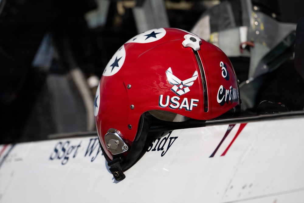 A red USAF flight helmet is resting on what appears to be the edge of a military aircraft cockpit. The helmet features the United States Air Force insignia, the text 'USAF', the number '3', and partial text reading 'Crim' (likely 'Crimson'). The aircraft surface below shows partial text reading 'SSgt Wy...dy', indicating a crew member's name.