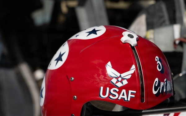 A red USAF flight helmet is resting on what appears to be the edge of a military aircraft cockpit. The helmet features the United States Air Force insignia, the text 'USAF', the number '3', and partial text reading 'Crim' (likely 'Crimson'). The aircraft surface below shows partial text reading 'SSgt Wy...dy', indicating a crew member's name.
