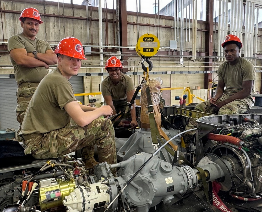 Four military personnel wearing red hard hats and olive/camouflage uniforms are working on what appears to be a helicopter transmission or gearbox assembly in an industrial maintenance hangar. A Yale 3-ton chain hoist is visible overhead, suggesting heavy component lifting is in progress. The team appears to be posing briefly for the photo during an aircraft maintenance operation.