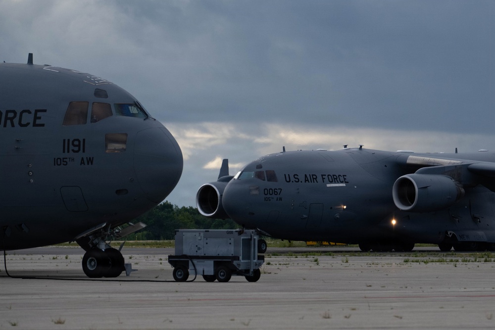 Two U.S. Air Force C-17 Globemaster III aircraft are parked on a flight line ramp under overcast skies. Both aircraft belong to the 105th Airlift Wing, with tail numbers 1191 and 0067 visible. Ground support equipment is positioned between the aircraft on the tarmac.
