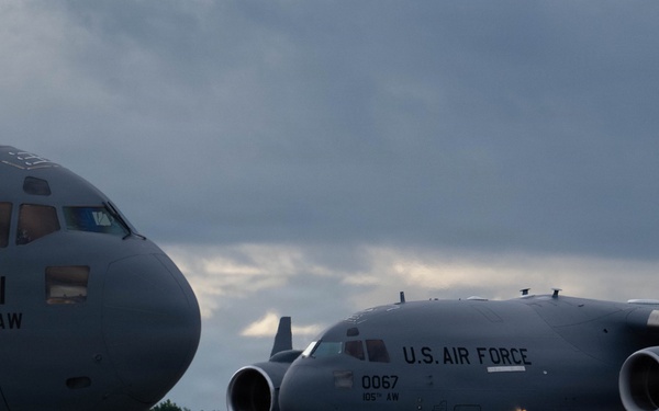 Two U.S. Air Force C-17 Globemaster III aircraft are parked on a flight line ramp under overcast skies. Both aircraft belong to the 105th Airlift Wing, with tail numbers 1191 and 0067 visible. Ground support equipment is positioned between the aircraft on the tarmac.