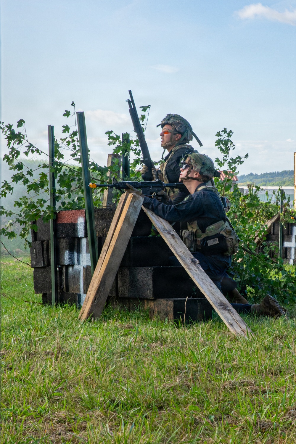 Two individuals in tactical military-style gear are positioned behind a makeshift cover constructed from concrete blocks and wooden planks in an outdoor field setting. Both are wearing helmets and plate carriers, with one aiming a rifle while the other observes. The setting and equipment suggest a military training exercise or airsoft/milsim event.