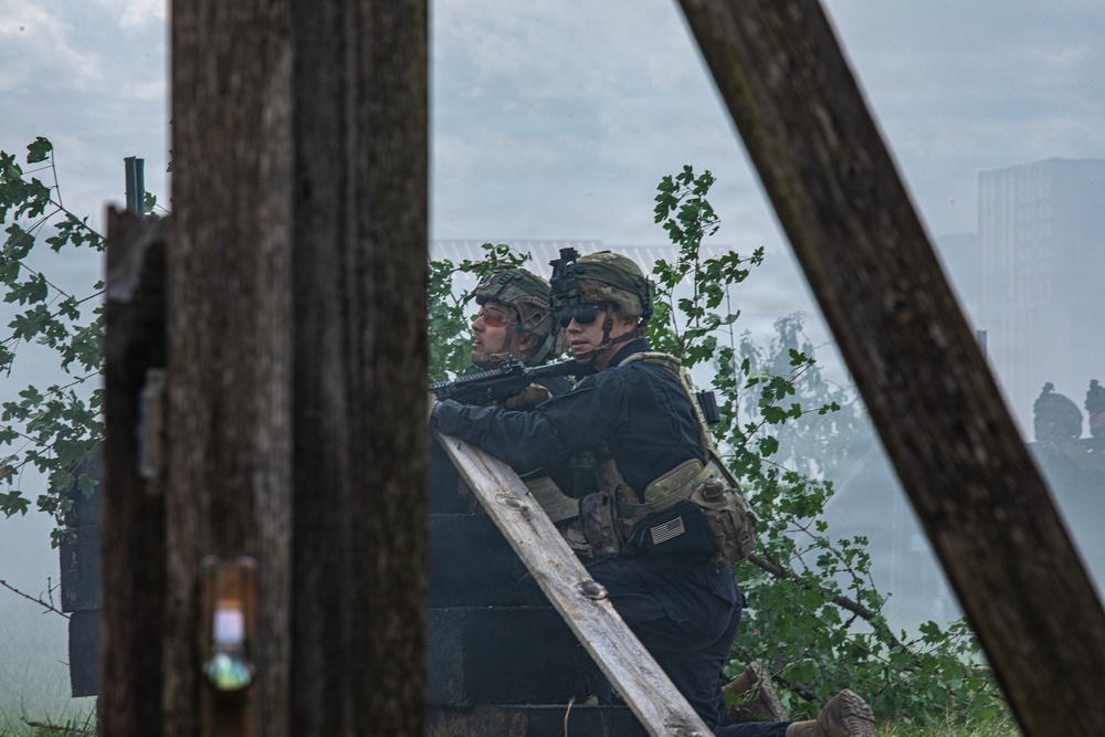 Two armed military personnel in full tactical gear are taking cover behind a low concrete or stone wall, scanning an area ahead. They are wearing camouflage helmets, tactical vests, and are equipped with rifles. The scene appears to be a military training exercise, evidenced by smoke in the background and what appears to be a training range environment.