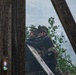 Two armed military personnel in full tactical gear are taking cover behind a low concrete or stone wall, scanning an area ahead. They are wearing camouflage helmets, tactical vests, and are equipped with rifles. The scene appears to be a military training exercise, evidenced by smoke in the background and what appears to be a training range environment.