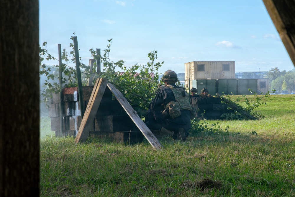 The image shows military personnel in tactical gear taking cover behind makeshift wooden barriers and vegetation in a grassy field. The scene appears to be a military training exercise, with soldiers in camouflage uniforms and helmets in low crouching positions. Modular military-style buildings or containers are visible in the background.