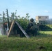 The image shows military personnel in tactical gear taking cover behind makeshift wooden barriers and vegetation in a grassy field. The scene appears to be a military training exercise, with soldiers in camouflage uniforms and helmets in low crouching positions. Modular military-style buildings or containers are visible in the background.