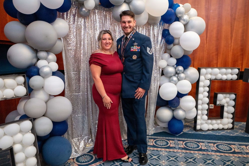 Two individuals pose for a formal photo at what appears to be a military ball or formal gala event. The man is wearing a U.S. Air Force dress uniform with multiple medals and ribbons, while the woman is wearing an elegant red/burgundy form-fitting gown. They are standing in front of a decorative backdrop featuring a sequined curtain, a balloon arch in blue, white, and silver, and large marquee letter decorations.