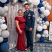 Two individuals pose for a formal photo at what appears to be a military ball or formal gala event. The man is wearing a U.S. Air Force dress uniform with multiple medals and ribbons, while the woman is wearing an elegant red/burgundy form-fitting gown. They are standing in front of a decorative backdrop featuring a sequined curtain, a balloon arch in blue, white, and silver, and large marquee letter decorations.