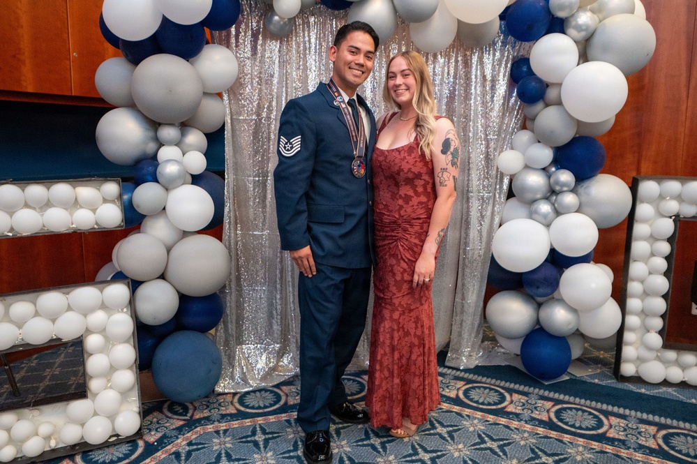 Two individuals pose together at a formal military event, likely an Air Force ball or awards ceremony. The man is wearing a US Air Force dress uniform with a Staff Sergeant rank insignia and a medallion/award around his neck. The woman is wearing a red/burgundy formal gown and has visible tattoos on her arm.