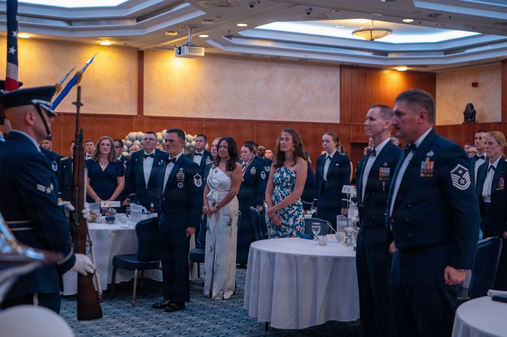 A formal military banquet or dining-in ceremony is taking place in a large ballroom or event hall. Military personnel in dress uniforms (dark blue with bow ties and medals) and civilian guests are standing at attention as a color guard presents flags at the front of the room. The setting includes round banquet tables with white tablecloths, suggesting a formal ceremonial dinner event.
