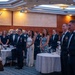 A formal military banquet or dining-in ceremony is taking place in a large ballroom or event hall. Military personnel in dress uniforms (dark blue with bow ties and medals) and civilian guests are standing at attention as a color guard presents flags at the front of the room. The setting includes round banquet tables with white tablecloths, suggesting a formal ceremonial dinner event.