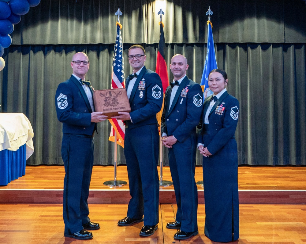 Four U.S. Air Force personnel in dress blue uniforms are posed on a stage during what appears to be a formal military ceremony or awards banquet. Two individuals in the center are jointly holding a wooden engraved plaque bearing the name 'JASON TRICKEY.' The setting includes U.S., German, and U.S. Air Force flags, blue balloons, and a decorated stage backdrop.