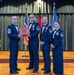 Four U.S. Air Force personnel in dress blue uniforms are posed on a stage during what appears to be a formal military ceremony or awards banquet. Two individuals in the center are jointly holding a wooden engraved plaque bearing the name 'JASON TRICKEY.' The setting includes U.S., German, and U.S. Air Force flags, blue balloons, and a decorated stage backdrop.